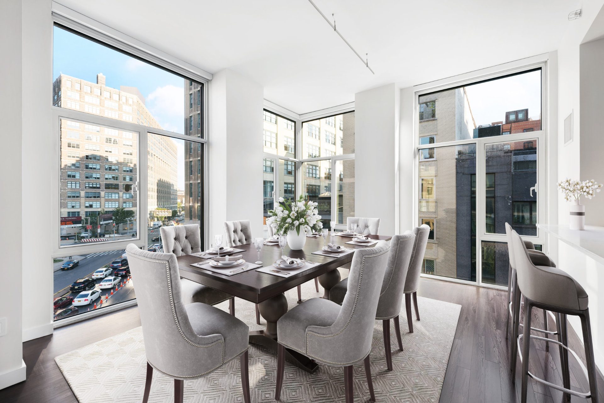 Tribeca II — dining room with a set table and upholstered chairs beside tall city-facing windows, with white built-in shelving and dark hardwood flooring
